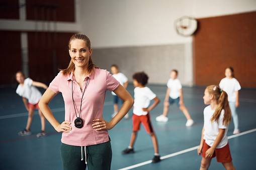 Portrait of happy female physical education teacher at school gym looking at camera. Students are exercising in the background.