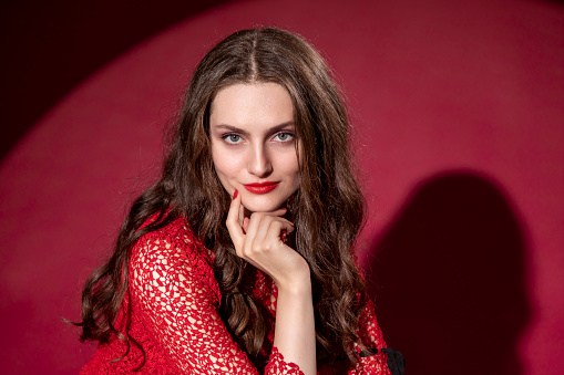 Portrait of a young woman with long brown hair in a red dress on a red background. She is looking at camera