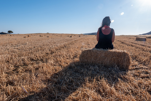A woman sitting on a bale of straw in a mown wheat field and watching the sunset. A wide angle landscape with selective focus.