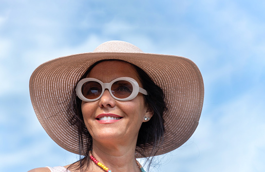 Woman with hat and sunglasses, smilling, on a summer day, with blue sky in the background.