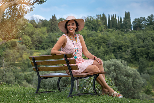 Mature gorgeous woman sitting on a bench on a top of the hill with trees  and clear sky in the background.