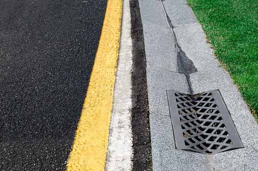 gutter of a stormwater drainage system on the side of an road with yellow and white markings and green lawn on wet weather after rain, close up view, nobody.