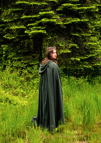Brunette young adult girl looking over her shoulder in green cloak and jeweled crown against a forest background.