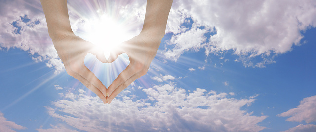 blue sky and fluffy clouds with a beautiful bright sun above female hands making a heart shape and copy space