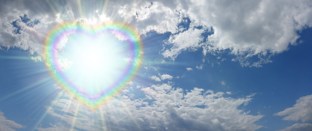 blue sky and fluffy clouds with a beautiful bright sun peering through a rainbow coloured heart frame with copy space