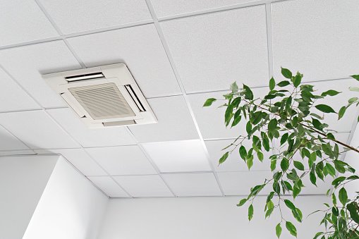 Low angle of assette Air Conditioner on ceiling in modern light office or apartment with green ficus plant leaves. Indoor air quality