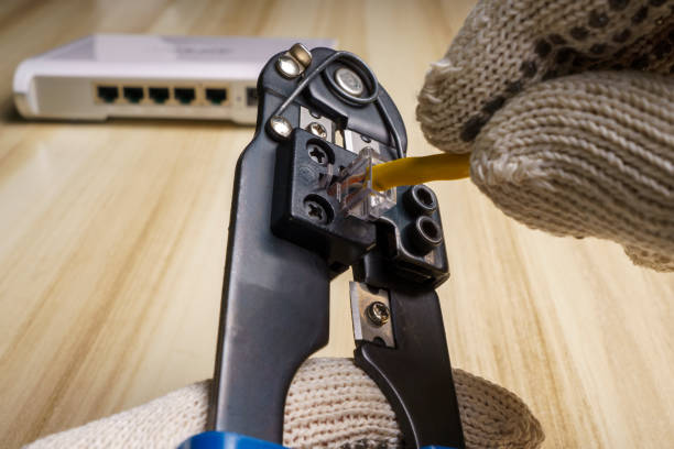 Gloved hands assembling a network connector with a tool for this purpose. Router out of focus from behind.