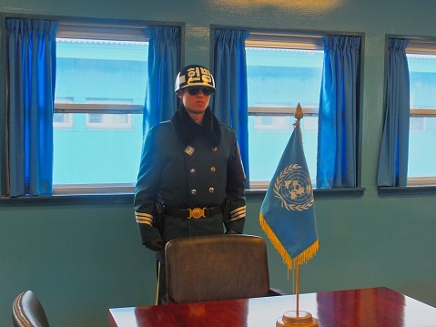 Panmunjom, South Korea – November 30, 2013: A South Korean soldier stands guard on the exact border between North and South Korea. The border is denoted by the row of microphones and the United Nations flag. The two countries have been at war since 1950. The border between them - known as the Demilitarized Zone, DMZ, or Joint Security Area, JSA - is the most heavily guarded border in the world. Here, in the Blue House, officials from North and South Korea can meet without officially leaving their countries.