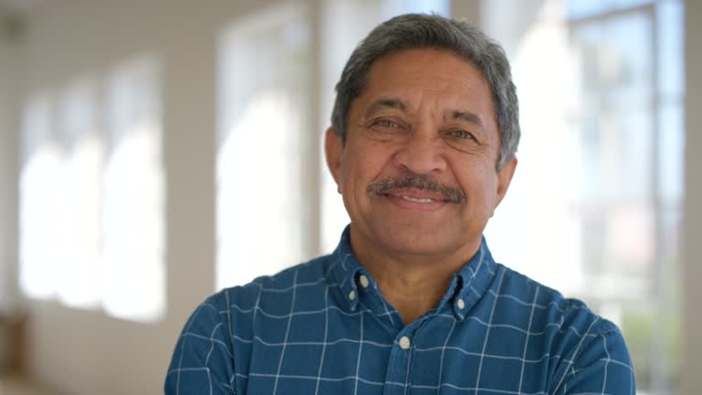 Portrait of a happy retired male looking cheerful standing inside a bright room. Face of a laughing mature latino man standing in an empty office or home. Smiling confident senior enjoying retirement