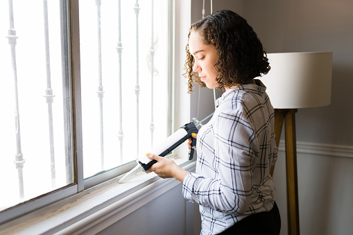 Attractive young woman using silicone sealant on the window and repairing the windows of her house