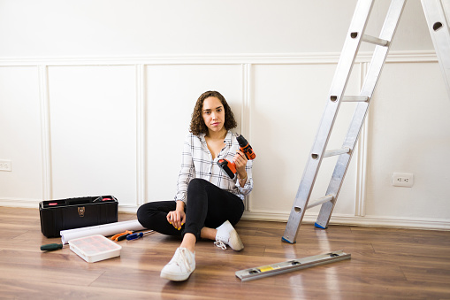 Portrait of a handy young woman looking at the camera while using a drill and a ladder to home repairs