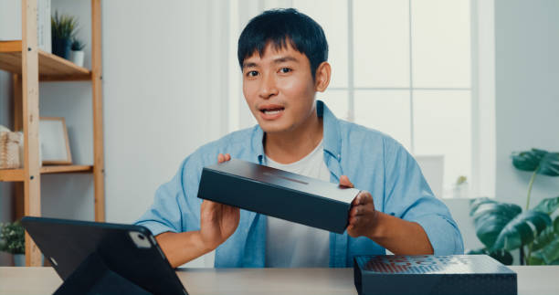 Young man blogger opening parcel box and recording unboxing video with camera sitting at work desk in living room at home.