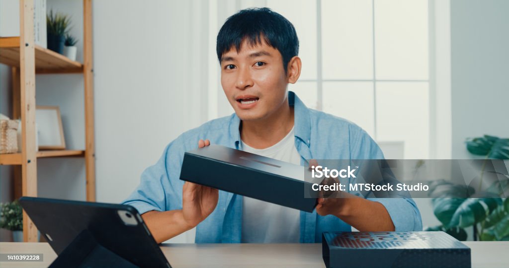 Young man blogger opening parcel box and recording unboxing video with camera sitting at work desk in living room at home. Young Asian man blogger opening parcel box and recording unboxing video with camera sitting at work desk in living room at home. Merchandise Stock Photo