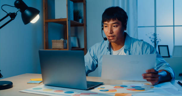 istock Young man wear headphone using laptop making video call with customer sitting at work desk in living room at home at night. 1410392499
