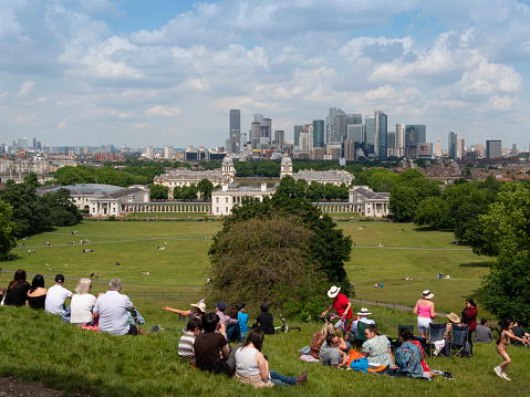 Groups of people sitting on the hill overlooking Greenwich and Canary Wharf in London Docklands on a fine day in June. They are awaiting the celebratory military flypast which is due to take place in an hour or so for the Platinum Jubilee of Queen Elizabeth II, which will be visible from Greenwich Park.