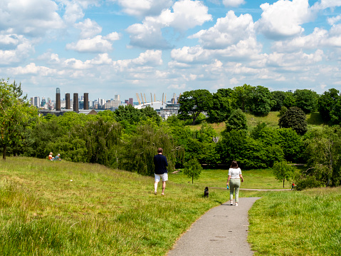 A woman and man walking their dog down a pathway in Greenwich Park, south east London. The chimneys of Greenwich Power Station can be seen with part of London Docklands on the other side of the River Thames and the O2 (former Millennium Dome) on the Greenwich Peninsula.