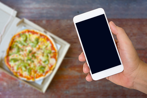 Man hand holding smartphone with blur image of pizza box on wood table background. Food delivery online concept.