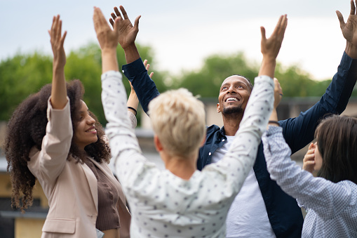 A diverse group of people with their arms raised in the air as they stand in a circle outside under the sky