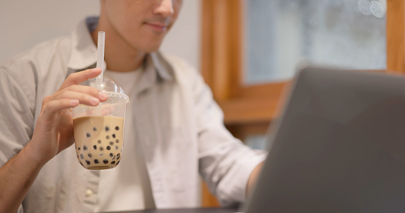 Asian young man works with laptop and drinks Bubble milk tea at cafe - popular food and drink in Taiwan