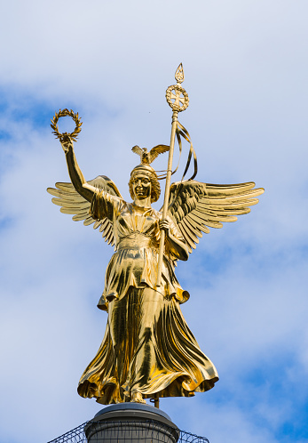 The golden statue on top of the victory Column (Siegessäule) in front of the blue midday sky and some clouds.