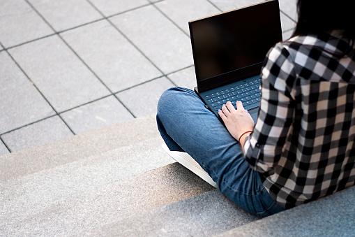 A woman sits on the steps in a black and white checkered shirt outside and holds a black laptop on her lap. Concept. Close-up. soft focus. Back view.