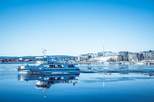 Oslo, Norway - March 6, 2022: A small ferry crossing the frozen Oslo Fjord in winter.  The famous Aker Brygge district in the background.