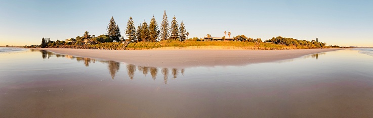 Horizontal landscape panoramic view of the beach, foreshore, coastal vegetation and pine trees reflecting on the wet sand at Belongil Beach, Byron Bay, north coast NSW in Winter.
