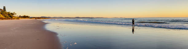 Horizontal landscape panorama of a man fishing, in silhouette, on Belongil Beach, Byron Bay, NSW, as the sun rises over the Pacific Ocean in Winter.