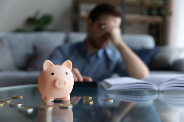 Young man feeling stressed calculating expenditures or taxes. Close up focus on piggybank on table, young man feeling stressed calculating expenditures or taxes, managing future payments or planning investments, suffering from lack of money, accounting concept. cost of living stock pictures, royalty-free photos & images