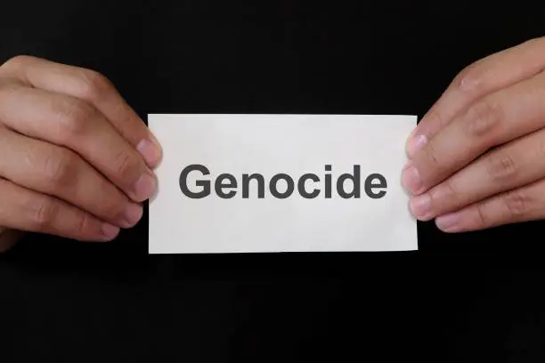 Genocide crime case mug shot concept. Criminal hands holding paper placard with written word in dark black background.