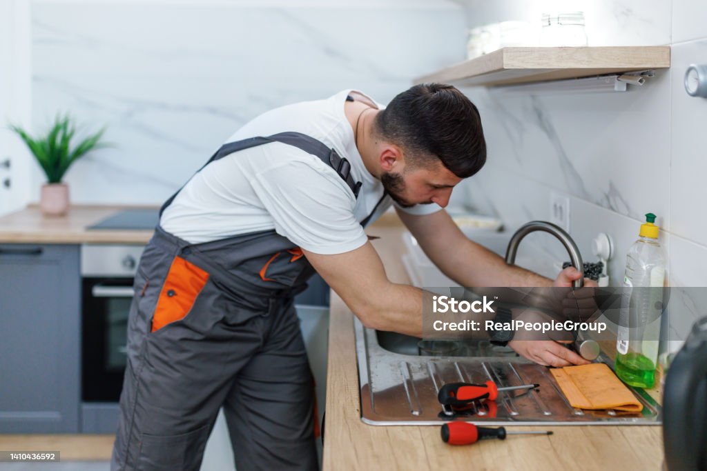 This faucet is leaking again A young Caucasian plumber is trying to tighten a leaking faucet on a kitchen sink. Repairing Stock Photo