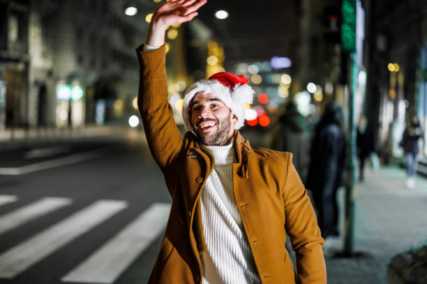 サンタの帽子をかぶった若い男性が通りに立っていて、タクシーを捕まえるために手を振っています。 - christmas fashion model human arm beautiful ストックフォトと画像