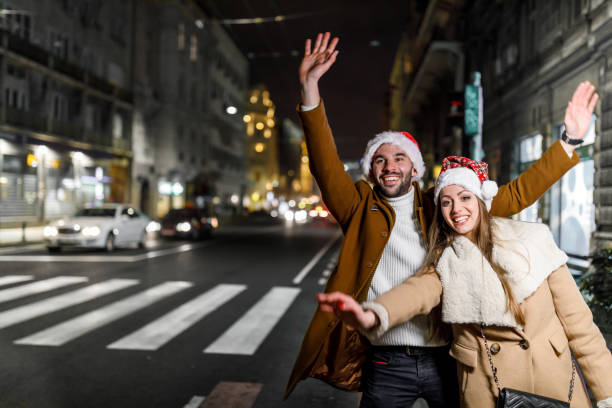 un jeune couple avec des chapeaux de père noël se tient dans la rue et agite le bras pour prendre un taxi. - christmas fashion model human arm beautiful photos et images de collection