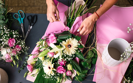 people, gardening and floral design concept - top down close up of woman or floral artist making bunch of flowers and tying bow on table at studio