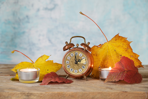 Switching from summer to winter time, vintage alarm clock showing the set back of one hour, autumn leaves and candles on rustic wood, light blue background, copy space, selected focus