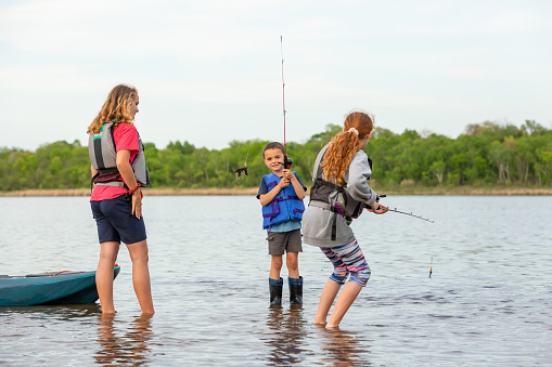 Two sisters and their younger brother fishing in the shallow water of a lake on a beautiful summer evening. The boy is showing off the leaf he caught.
