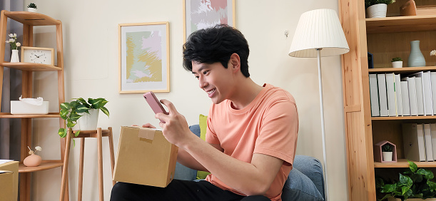 asian man look package use phone confirm information on parcel from online shopping delivery service sit sofa at home