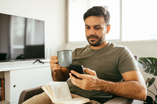 Young man using cellphone and drinking coffee or tea with after reading a book at home.