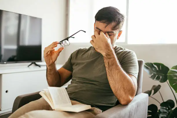 Photo of Tired man takes off his glasses and rubs his eyes while reading a book at home.