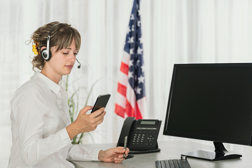 Caucasian female worker in headphones using mobile phone with blank screen computer with United States flag in background
