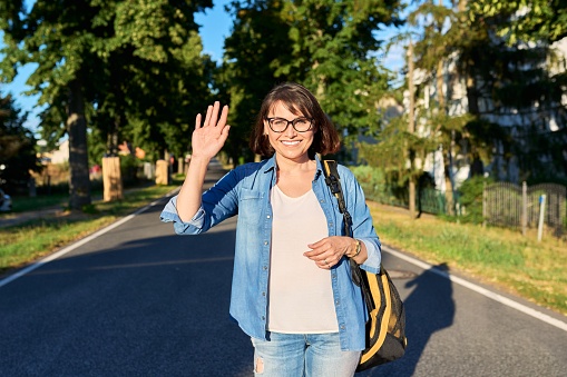 Happy middle aged woman with backpack waving hand looking at camera standing on the road in the street. Travel, tourism, journey, vacation, way, lifestyle, people 40s concept