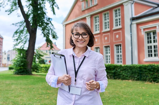 Middle-aged positive female school worker, teacher on background of school building. Woman psychologist advisor social worker with clipboard looking at camera outdoor. Education staff people concept