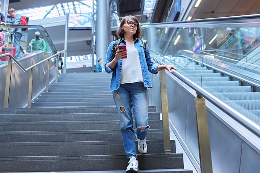 Woman with backpack walking up the stairs near escalator in modern station building. Urban architecture, city lifestyle, people concept