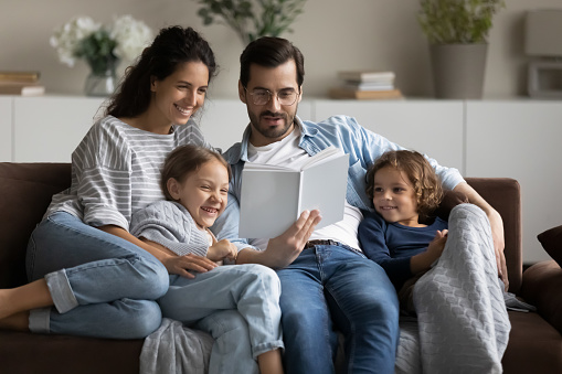 Young man his wife and cute preschool son and daughter reading book seated on sofa in living room, loving family spend leisure enjoy favourite hobby. Couple develop their kids, pastime at home concept