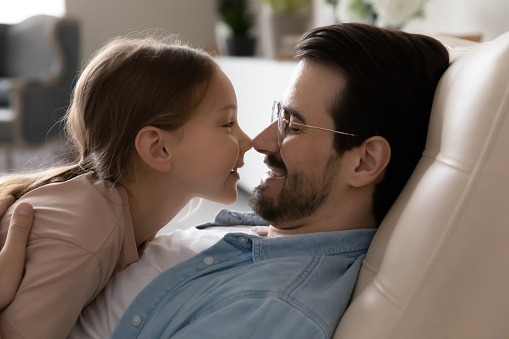 Close up faces of loving dad and cute 6s daughter rest on sofa touch noses looking at each other with love, enjoy moment of tenderness, spend free time together at home. Family bond and ties concept