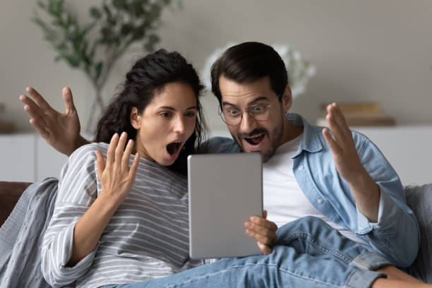 Astonished couple gawp at digital tablet feel amazed by news Astonished couple sit on sofa open mouth gawp at digital tablet screen feel amazed shocked by unbelievable commercial offer, sell-out, huge discounts. Lottery winners enjoy moment of victory concept gawp stock pictures, royalty-free photos & images