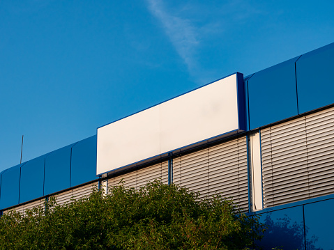 Blank name board on a building exterior. Empty white space for a company logo on a wall. Template of a signboard on an office house. Blue facade with closed window blinds. Blue sky in the background.