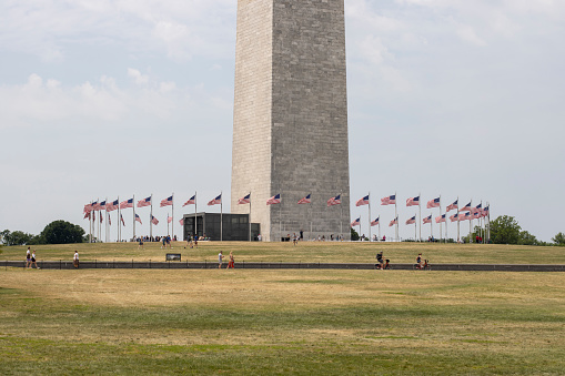 Washington, DC, USA - June 23, 2022: Visitors to the Washington Monument , an obelisk shaped building to commemorate George Washington, within the National Mall in Washington, DC, on a summer day. 50 American flags encircle the base of the monument.
