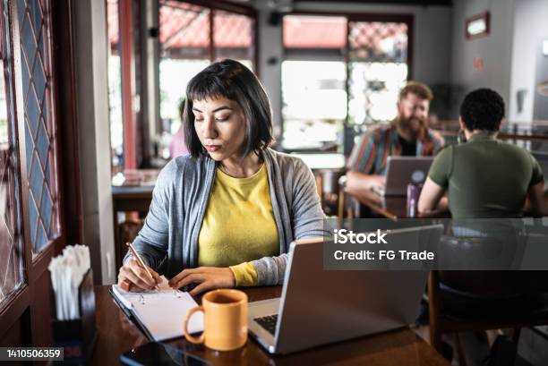Young Woman Writing On A Notepad And Using The Laptop In A Restaurant Stock Photo - Download Image Now