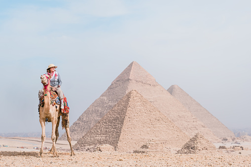 back view of tourist woman wearing a salacot riding a dromedary in front of pyramids. Egypt, Cairo - Giza